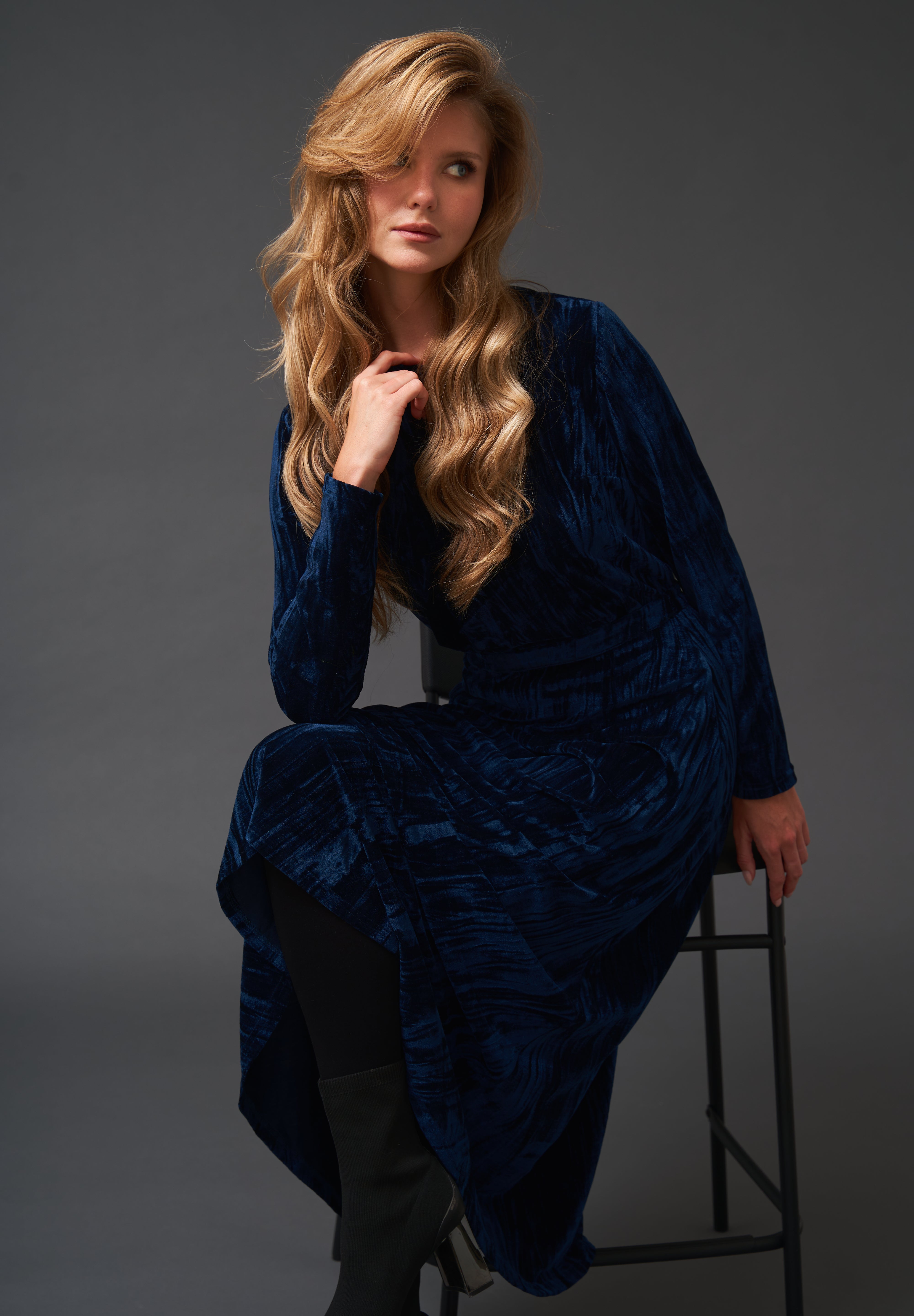 Woman wearing a blue velvet dress sitting on a stool against a gray background